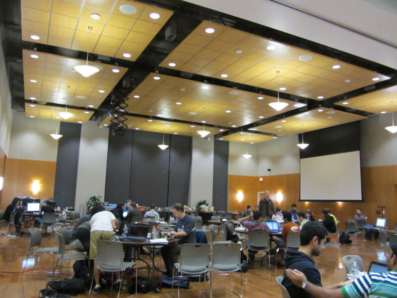 A wide hall with scattered tables of people working at laptops and a projector screen.