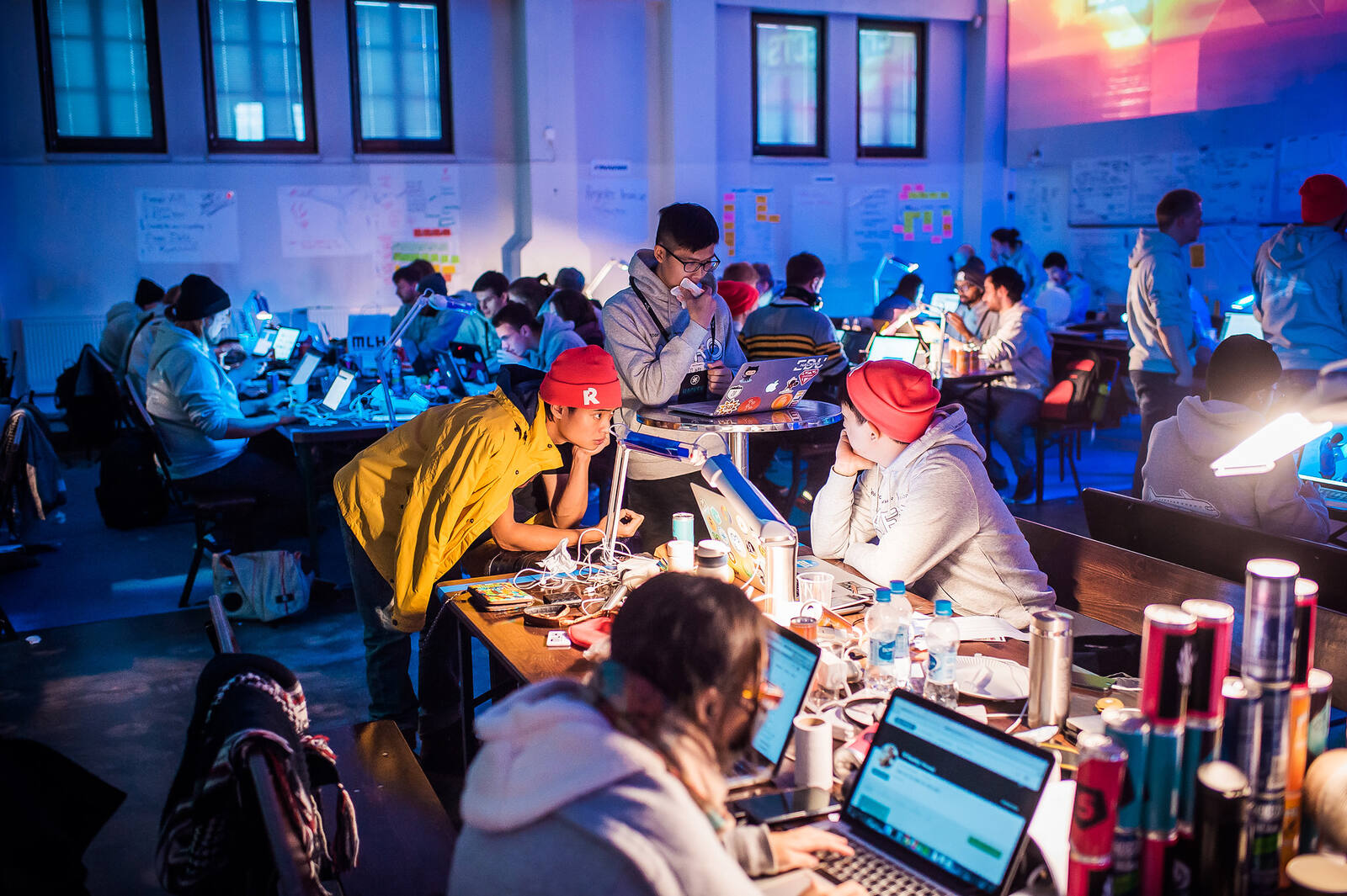 A hackathon floor at night: teams of builders at long tables lit by laptop screens and desk lamps, whiteboards covered in plans behind them.
